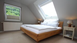 Ein Schlafzimmer mit schrägen Wänden, einem Bett mit weißer Bettwäsche, einem Nachttisch mit Lampe und einem Fenster mit Blick ins Grüne.