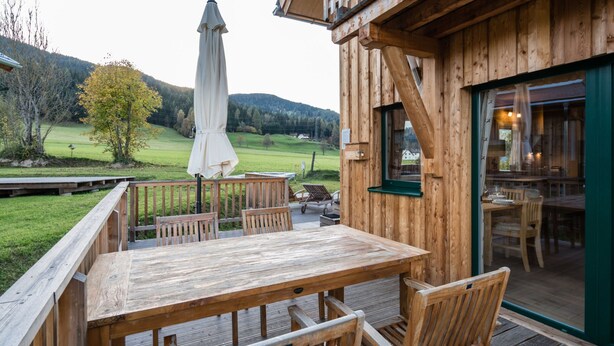 A wooden deck with a table and chairs, a covered umbrella, and a view of a green landscape with trees and hills in the background. - Amazing Chalet With hot tub (Sankt Georgen am Kreischberg)
