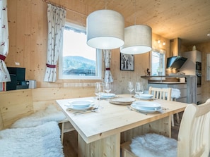 A wooden cabin interior with a dining table set for four, a kitchen area, and a window with a view of mountains. - Amazing Chalet With hot tub (Sankt Georgen am Kreischberg)