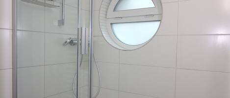 A modern bathroom with white tiles, a glass shower enclosure, a round mirror with a door, and a metal shelf.