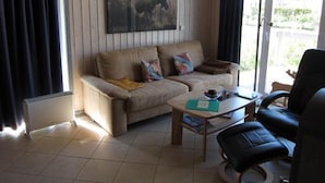 Un salón con sofá, una mesa de centro y una silla. Se aprecia una ventana con cortinas y un cuadro en la pared.