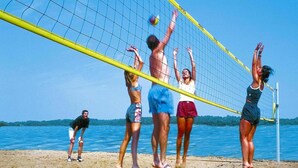 Een strandvolleybalwedstrijd met spelers in zwemkleding, een net en een zandstrand met water op de achtergrond.