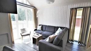 Een moderne woonkamer met een grijze bank, een kleine tafel en een stoel. Er is een groot raam met gordijnen en een plafondlamp.