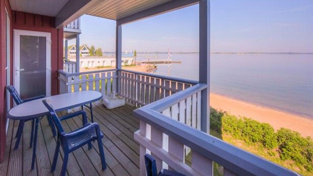 A wooden deck with a table and chairs, overlooking a calm body of water and a distant shoreline. - Exklusives Appartement in Rechlin (Rechlin)