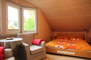Ein gemütliches Schlafzimmer im Dachgeschoss mit einem karierten Stuhl, einem kleinen runden Tisch und einem Bett mit orangefarbener Bettdecke.