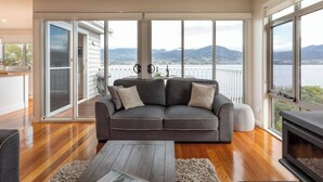Una sala de estar moderna con un sofá gris, suelo de madera y ventanas amplias que ofrecen vistas a un cuerpo de agua y montañas.