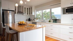 Una cocina moderna con armarios blancos, una isla de madera y un amplio ventanal con vistas a árboles y al mar.