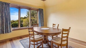 A dining area with a round wooden table and four chairs, large windows with curtains, and a view of greenery outside. - The Clocktower Serene Stay at Herons Rise Vineyard (Kettering)