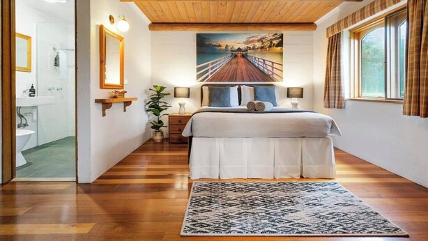 Un dormitorio con techo de madera, una cama grande, un cuadro de un muelle, una mesita de noche, una lámpara y una ventana con cortinas.