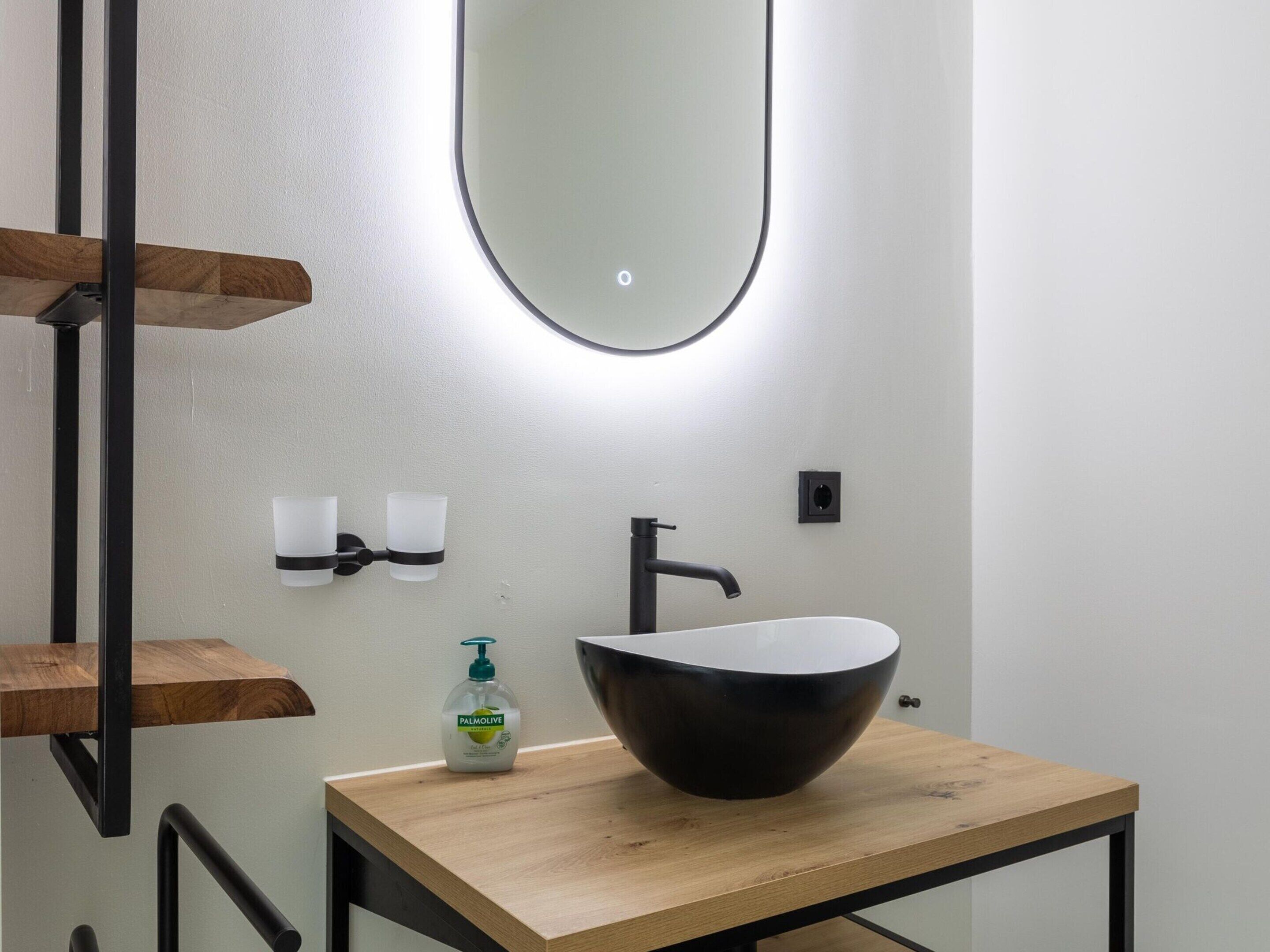 A modern bathroom with a wooden vanity, a large mirror, a black sink, and a bottle of Palmolive soap.