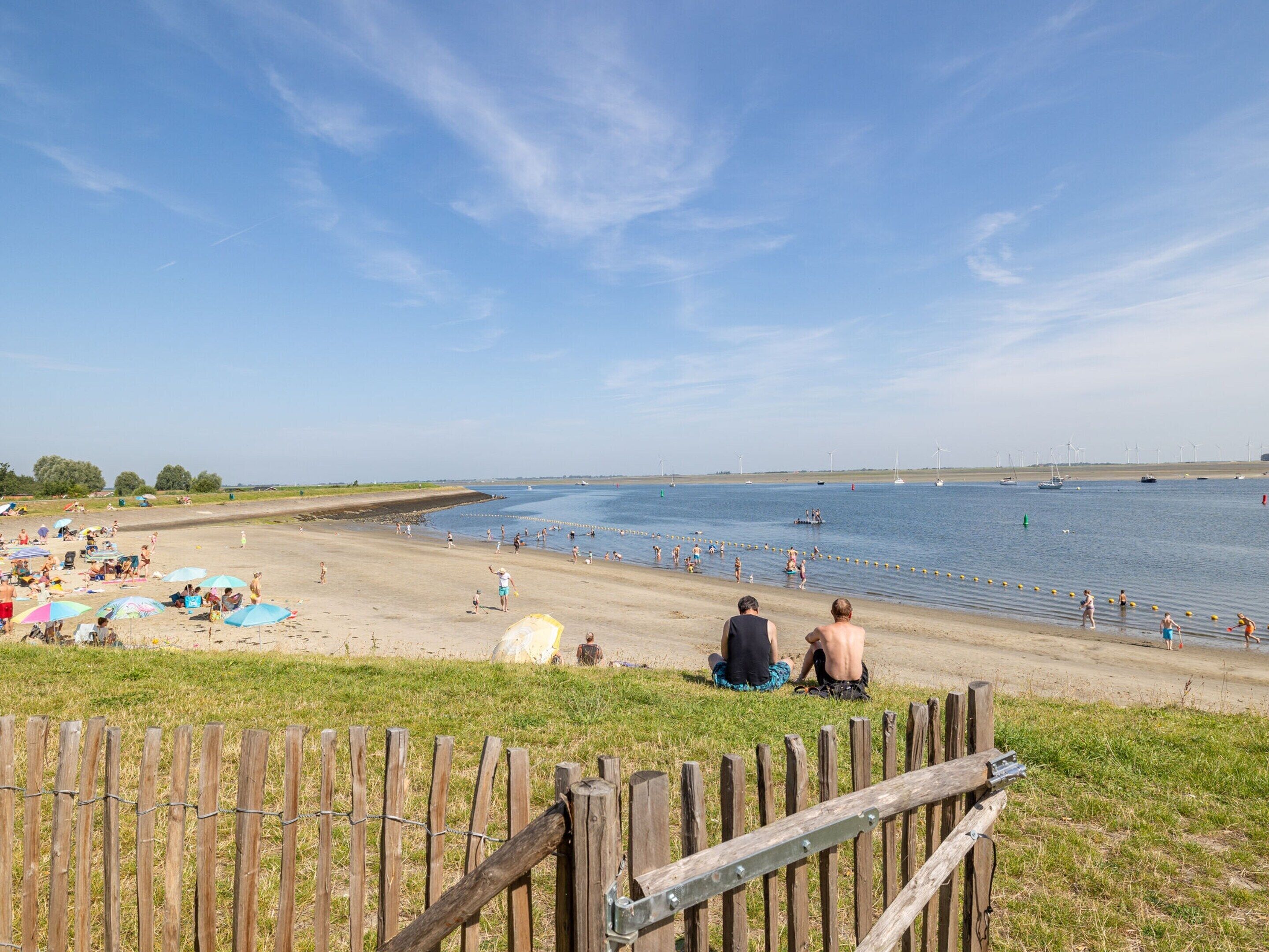 A beach with people sunbathing and swimming, a wooden fence in the foreground, and wind turbines in the distance.