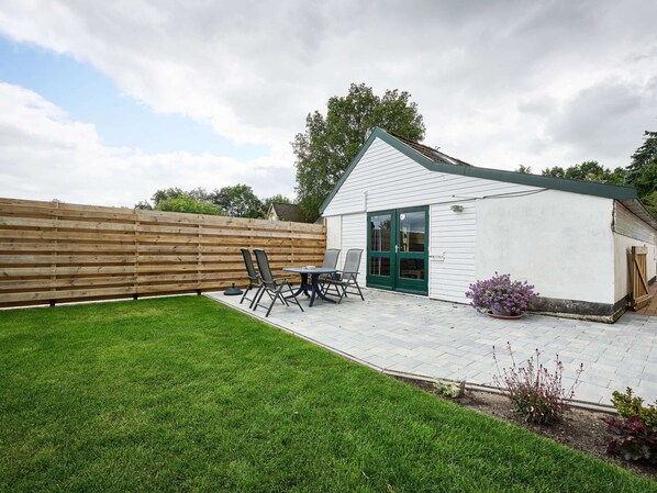 A small white building with a green door and windows, a patio with a table and chairs, and a wooden fence. - Cozy Holiday Home in a Lovely Environment (De Kiel)