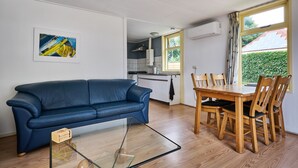 Una sala de estar con un sofá azul, una mesa de centro de vidrio y una mesa de comedor de madera con sillas. Se ve una cocina al fondo, con una ventana que da a la vegetación exterior.