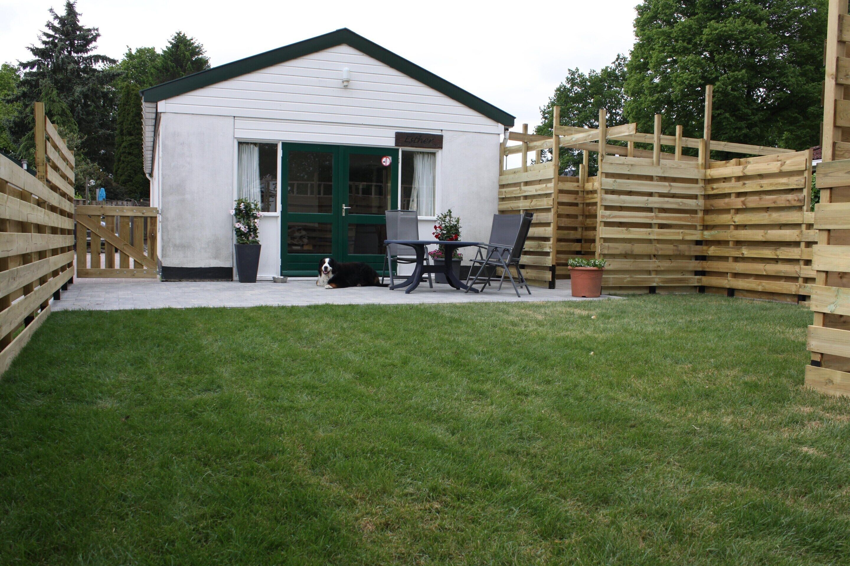 A small white building with a green door and windows, a wooden fence, a lawn, and two chairs and a table.