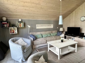 Una sala de estar con sofá, sillón, mesa de centro, estantería, televisor y un reloj en la pared.