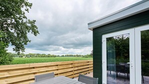 A wooden deck with a table and chairs, a glass door, and a view of a green field. - Stay at This Beautiful Accommodation in De Kiel (De Kiel)