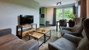 A modern living room with a flat-screen TV, wooden coffee table, and comfortable seating. A kitchen is visible in the background, and a balcony with a view of greenery is accessible through a sliding door. - Stay at This Beautiful Accommodation in De Kiel (De Kiel)