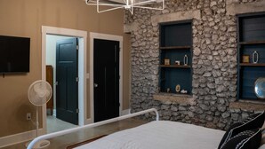 A bedroom with a stone wall, a bed with white bedding, a bedside table, a fan, and a TV mounted on the wall. - Historic 2 bedroom steps off downtown square (Mountain Home)