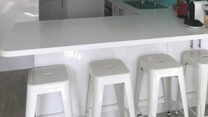 Eine moderne Küche mit weißer Arbeitsplatte, einer türkisblauen Spritzschutzwand und einer Edelstahl-Dunstabzugshaube. Drei weiße Hocker stehen an der Arbeitsfläche.