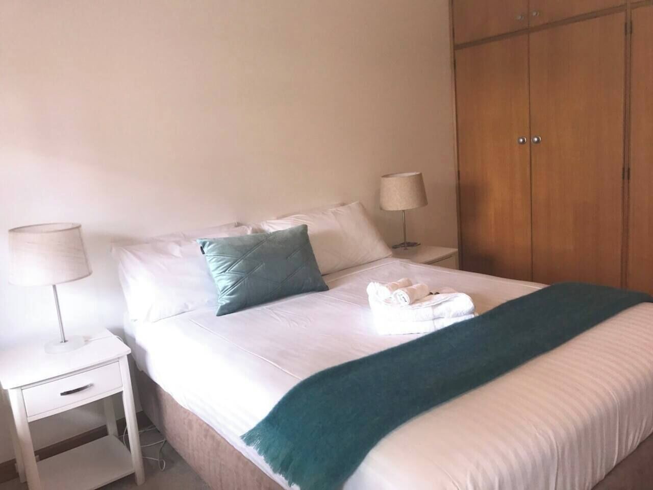Een net opgemaakt bed met witte lakens en een teal deken, een bedlampje en een houten garderobe.