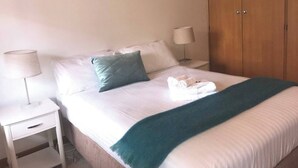 Ein ordentlich bezogenes Bett mit weißen Leinentüchern und einer türkisfarbenen Tagesdecke, eine Nachttischlampe und ein Holzschrank.