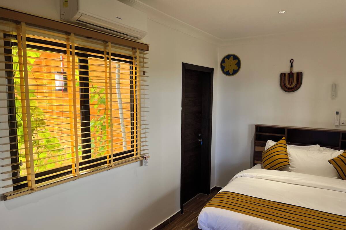 Quarto de hotel com uma cama, janela com persianas, porta e interruptor de luz fixado na parede.