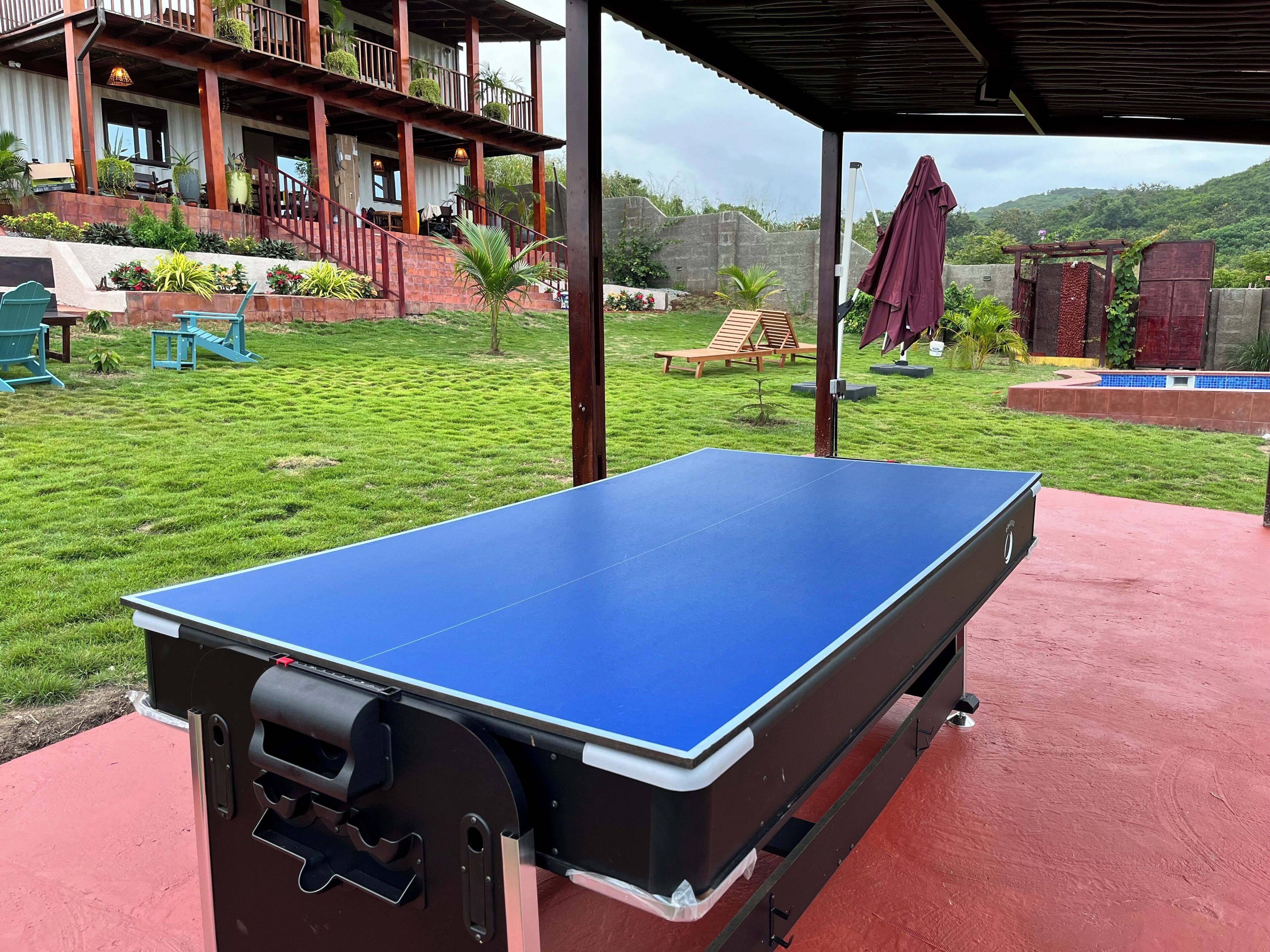 Ein Tischtennistisch steht im Freien unter einem Vordach, mit Blick auf ein Haus mit Balkon und Pflanzen.