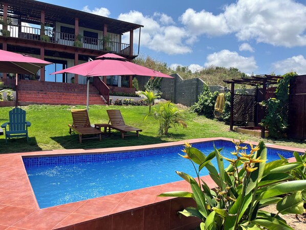 A swimming pool with a tiled edge, surrounded by a grassy area with lounge chairs and umbrellas.