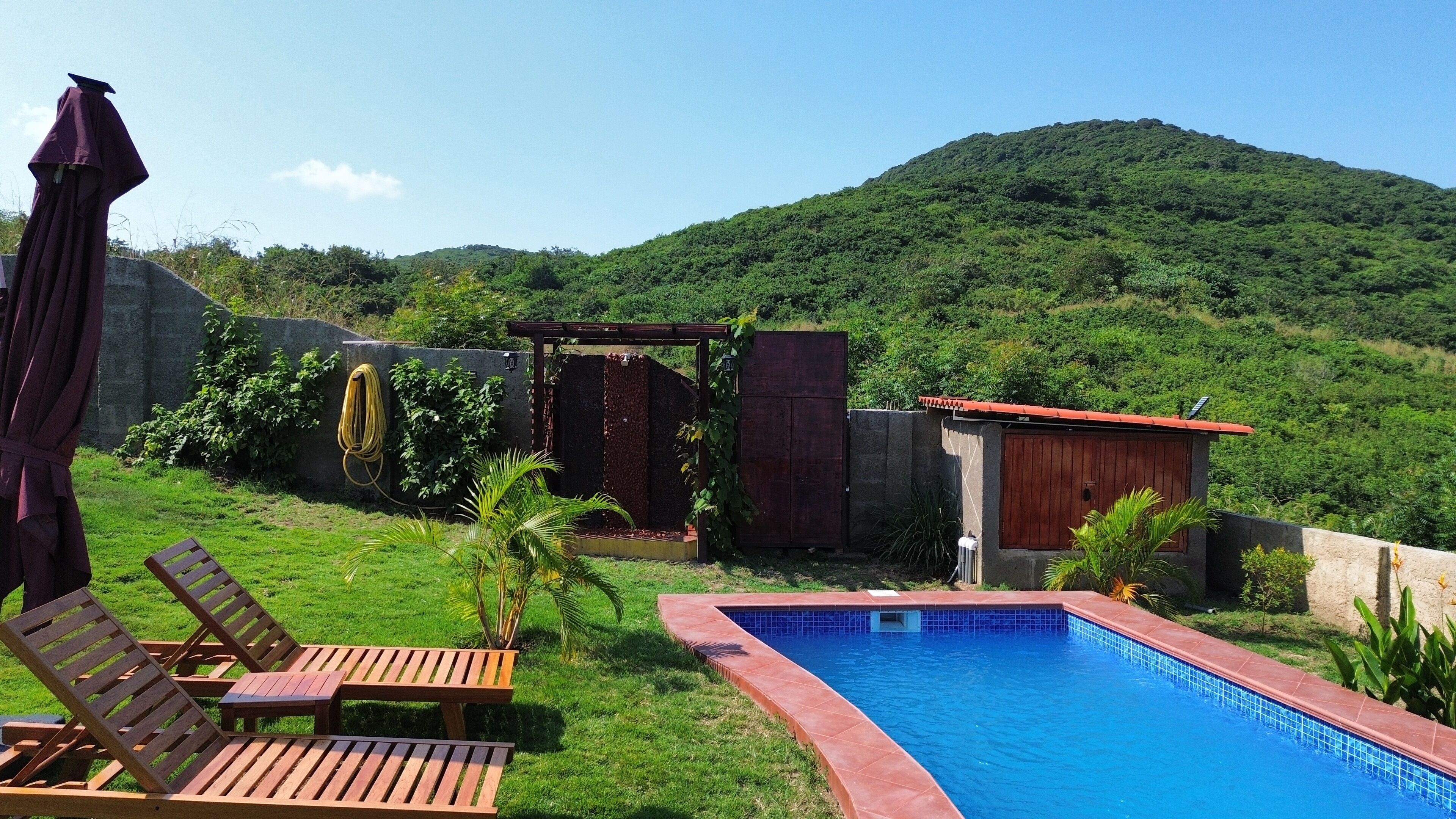 A backyard with a swimming pool, wooden lounge chairs, a pergola, and a view of a lush green hill.