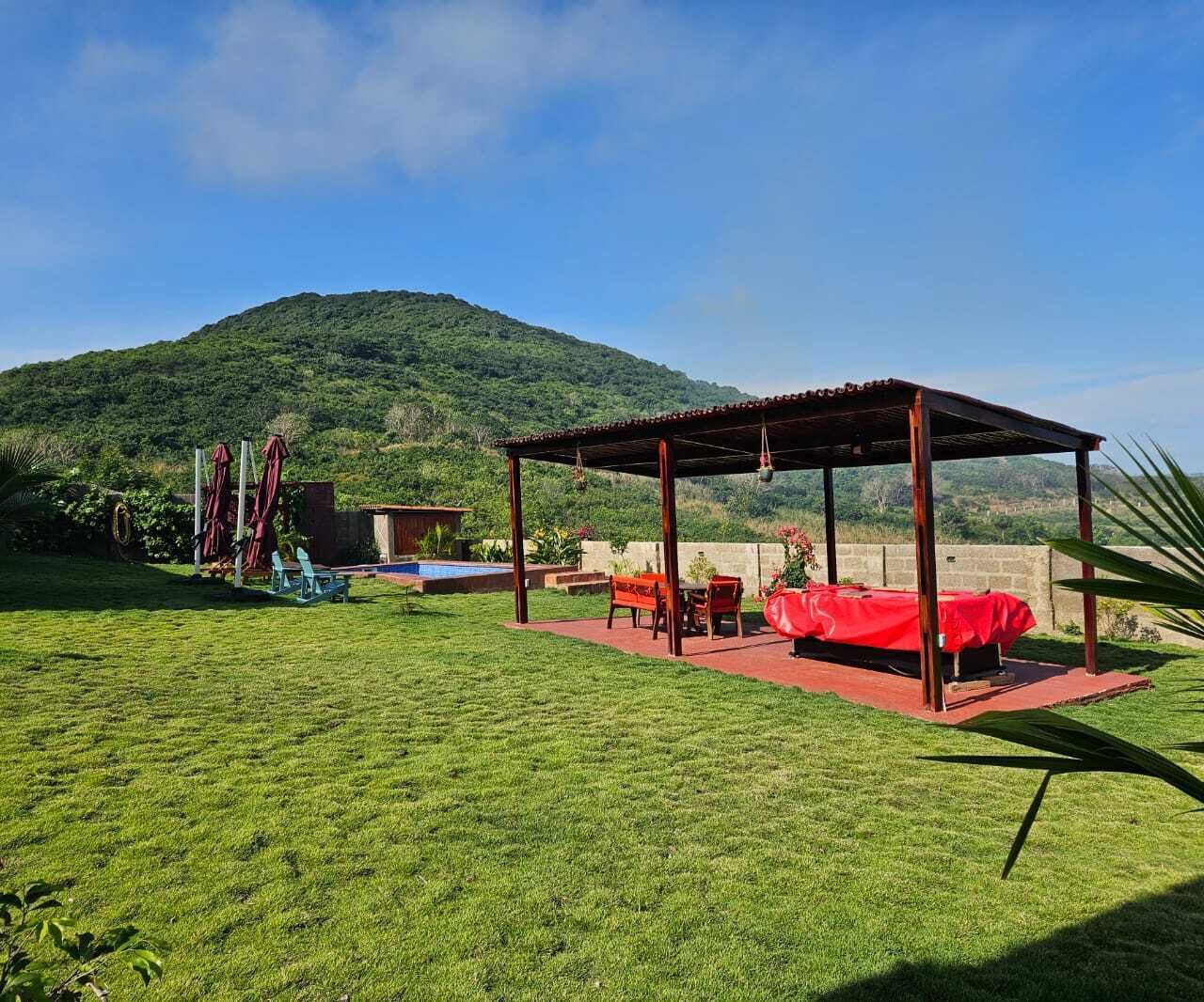A covered outdoor seating area with a red table and chairs, a small garden, and a mountain in the background.