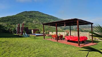 A covered outdoor seating area with a red table and chairs, a small garden, and a mountain in the background.