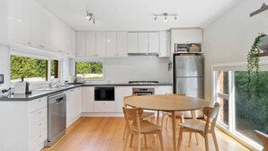 A modern kitchen with white cabinets, a stainless steel refrigerator, a built-in oven, and a dining area with a wooden table and chairs. - New Town Bungalow A Leafy Family Haven (New Town)