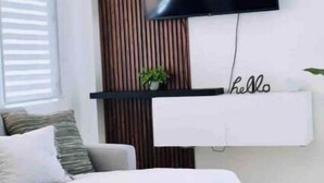A modern living room with a grey sofa, a mounted flat-screen TV, a wooden slat wall, and a minimalist shelf with a 'hello' sign. - Azores Luxury Residence

 (Borinquen)