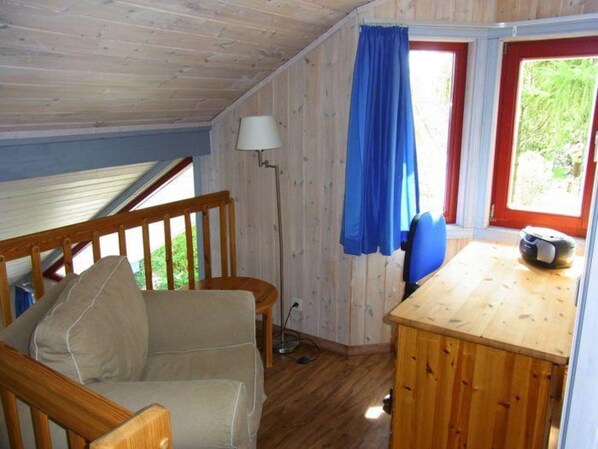 A room with a wooden ceiling, a wooden desk, a chair, a lamp, and a window with curtains.
