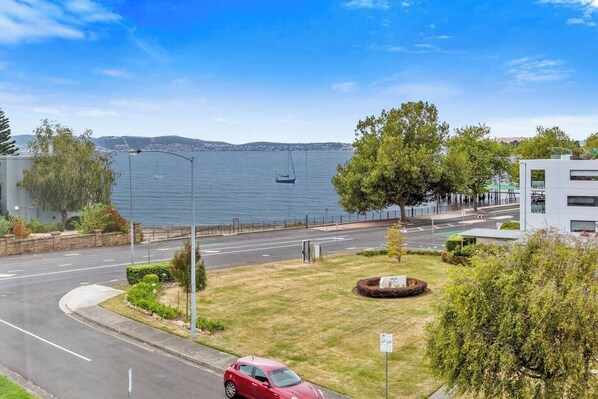 A coastal road with a red car parked on the side, a grassy area with a circular flower bed, and a view of the sea and distant hills.