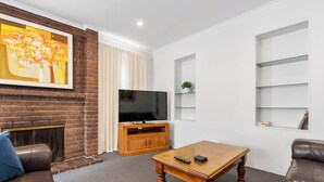 A modern living room with a brick fireplace, a flat-screen TV, a wooden coffee table, leather sofas, and a large abstract painting. - Classic Retro Home Steps from Sandy Bays Best (Sandy Bay)