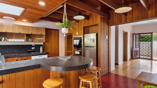 A modern kitchen with wooden cabinetry, a granite countertop, and a hanging plant. There are two wooden stools and a table with chairs. - Classic Retro Home Steps from Sandy Bays Best (Sandy Bay)