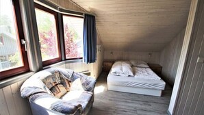 Een kleine zolderkamer met een enkel bed, een gebogen bankje en grote ramen.