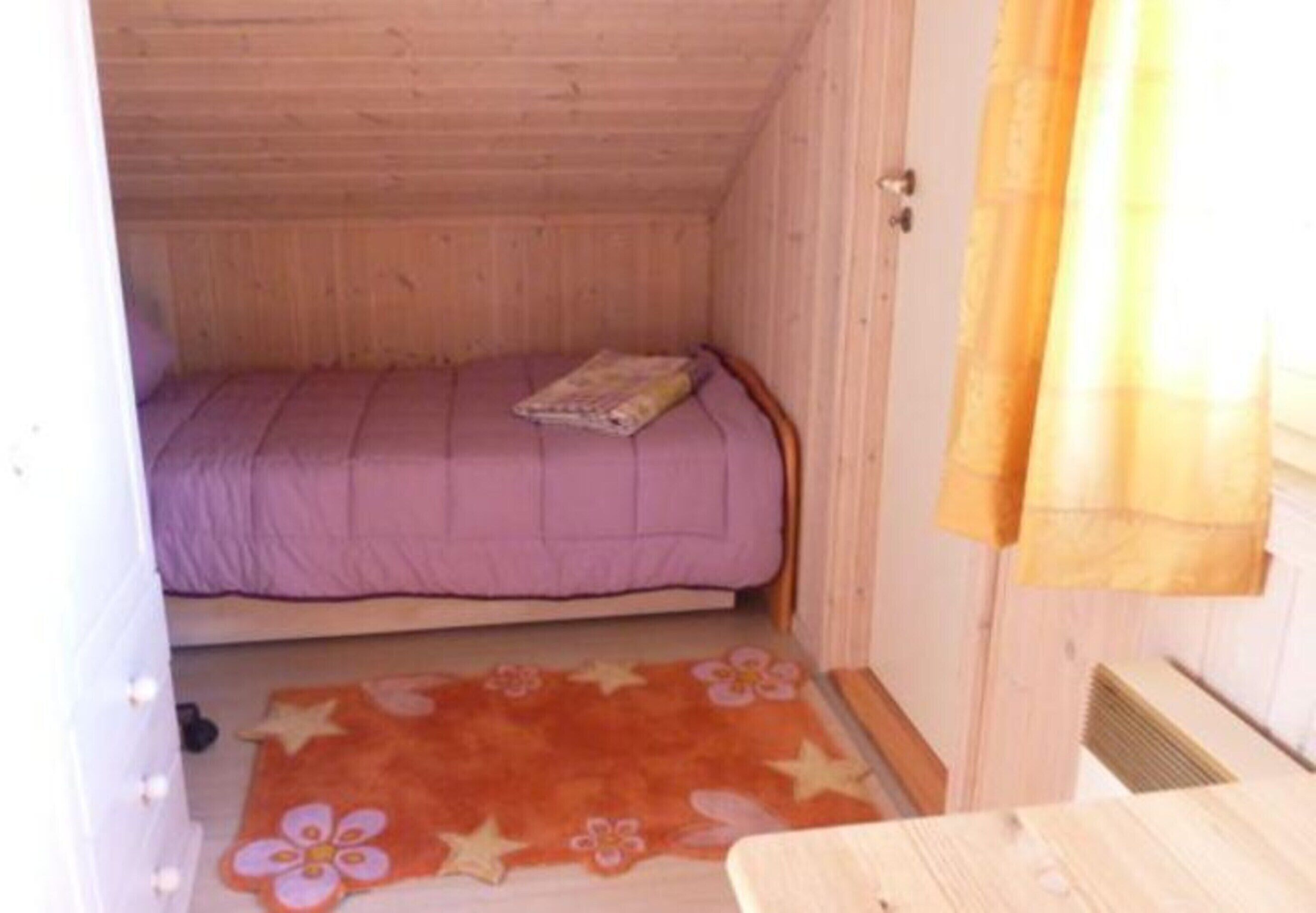 Ein kleines Zimmer mit einem Einzelbett, Holzboden und -wänden, einer violetten Tagesdecke, einem Blumenmuster-Teppich und einem kleinen Tisch.