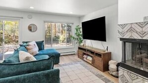 Een moderne woonkamer met een grote flatscreen televisie, een open haard en uitzicht op een bos door grote ramen.