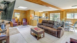 A cozy living room with a leather sofa, a coffee table, and a television. The room has wooden walls and a kitchen in the background. - 22 Mi to Lost Trail Skiing: Serene Winter Retreat (Sula)