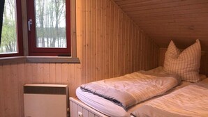 A small, cozy room with wooden walls and a sloped ceiling. There is a bed with pillows and a window with a view of trees. - Ferienhaus im Skandinavischen Stil (Mirow)