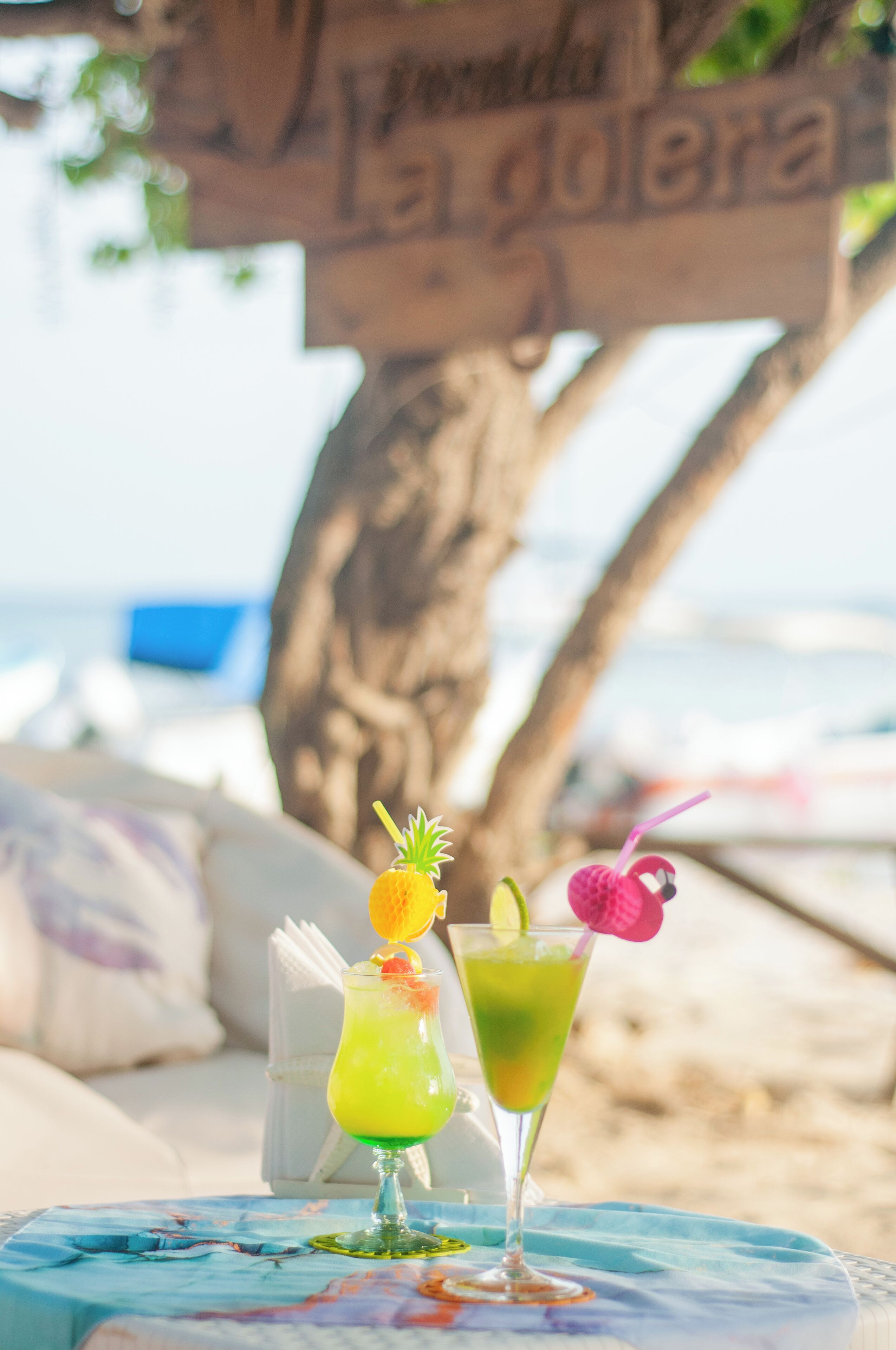 Dos cócteles sobre una mesa, con vista a la playa y un letrero de madera al fondo.