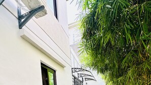 Een smal pad met witte kiezelstenen en groene beplanting, dat leidt naar een gebouw met een raam en een blauwe vat.