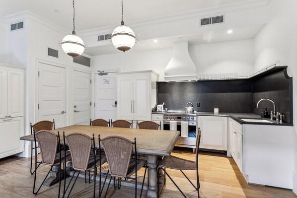 A modern kitchen with white cabinetry, a wooden dining table, wicker chairs, and a black countertop. - Hotel Perle (New Orleans)