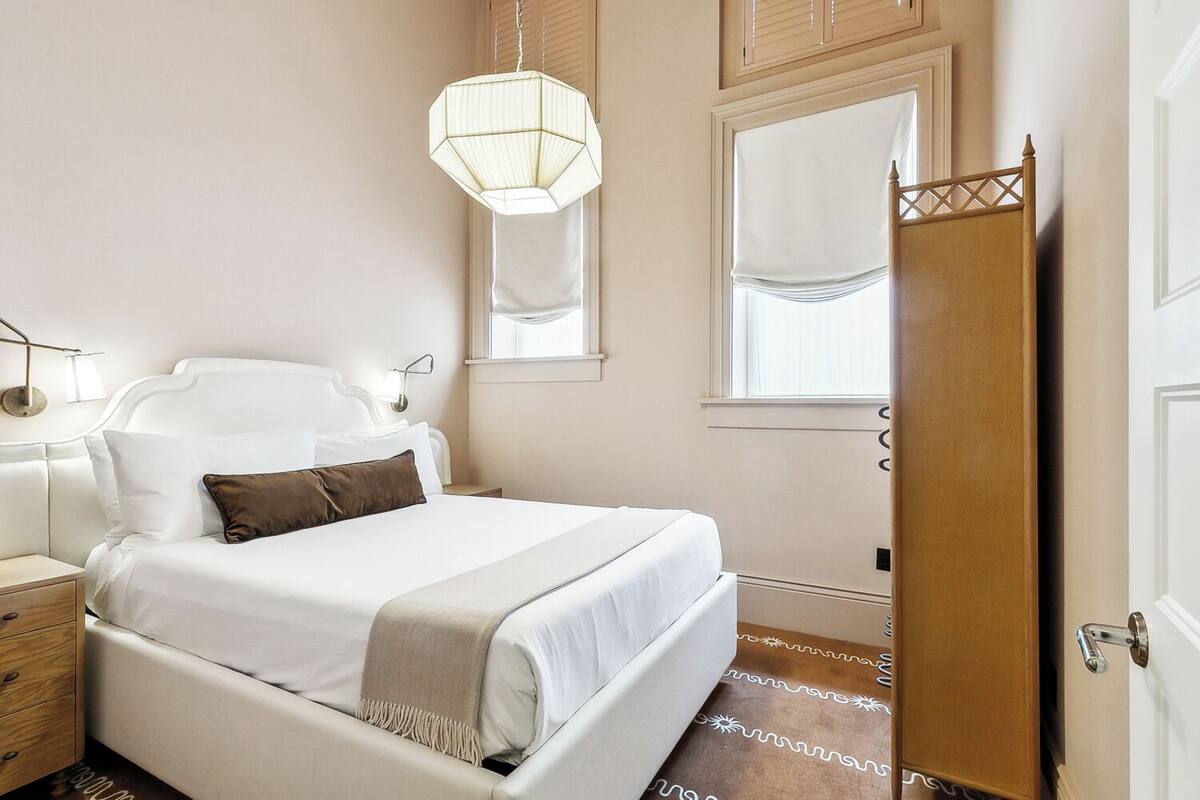 A bedroom with a bed, bedside tables, a hanging light fixture, a window with curtains, and a door.