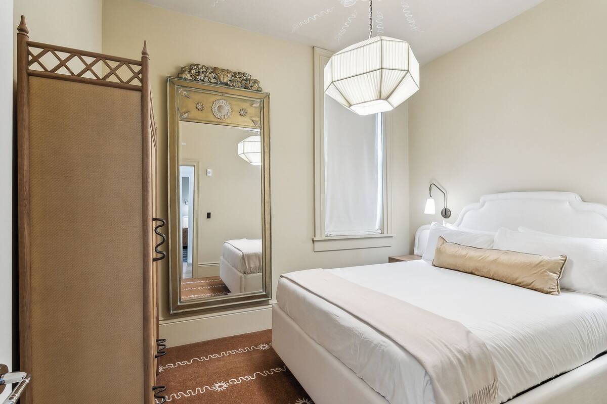 A bedroom with a large bed, a mirror, a window with curtains, and a hanging light fixture.