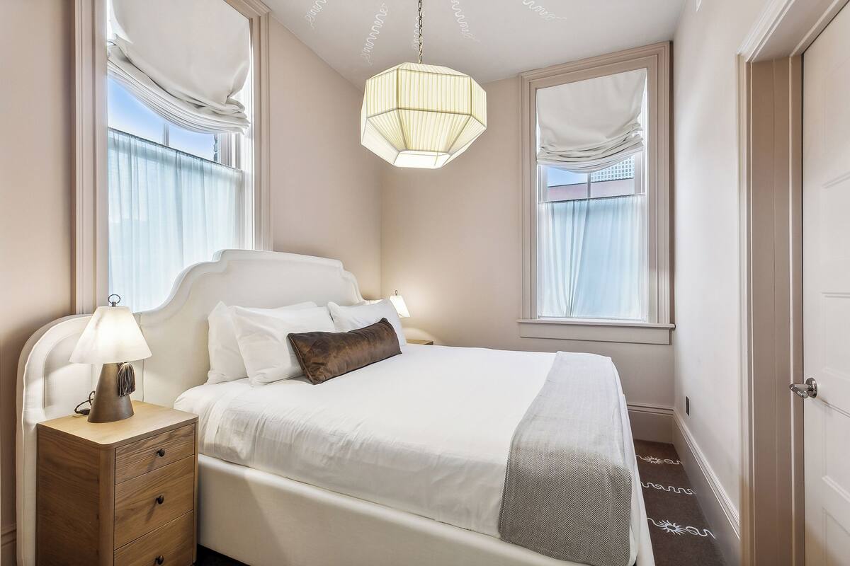 A neatly made bed with a white comforter and a brown pillow, a bedside table with a lamp, and a large window with curtains.