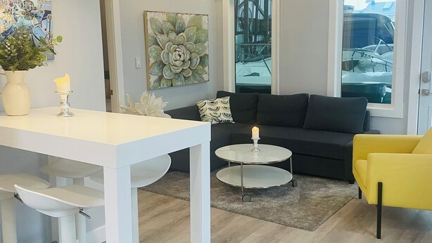 Un salón moderno con una mesa de comedor blanca, un sofá negro y un sillón amarillo. Hay un ventanal con vistas a barcos y un cuadro de flores en la pared.