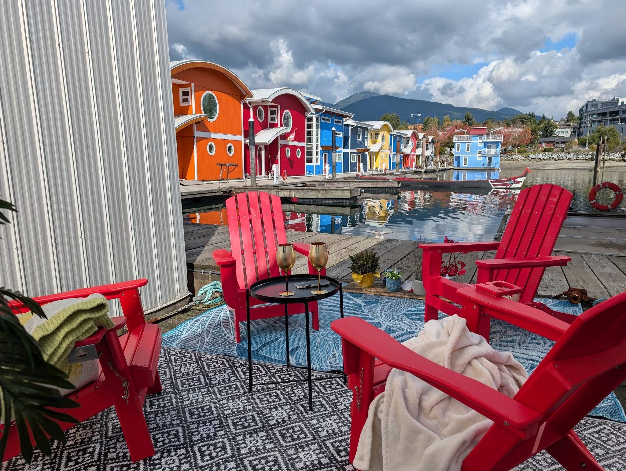 Eine Terrasse am Wasser mit roten Adirondack-Stühlen, einem kleinen runden Tisch mit Weingläsern und einem gemusterten Teppich.
